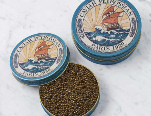 Une boîte de Caviar Petrossian Paris 1920 dont le couvercle est partiellement ouvert, révélant une masse de caviar noir à l'intérieur. Le couvercle, conçu graphiquement par Claire Duclert, présente une illustration d'un navire sur l'océan avec le nom de la marque et le logo, posés sur une surface en marbre.