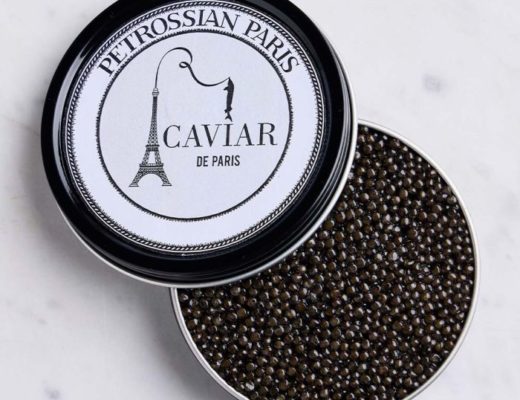 Une boîte ronde de caviar Petrossian Paris dont le couvercle est partiellement ouvert, révélant des perles de caviar noires à l'intérieur. Le couvercle, conçu par Claire Duclert, arbore le logo Petrossian avec une illustration de la Tour Eiffel et le texte « Caviar de Paris ». La boîte repose sur une surface en marbre.