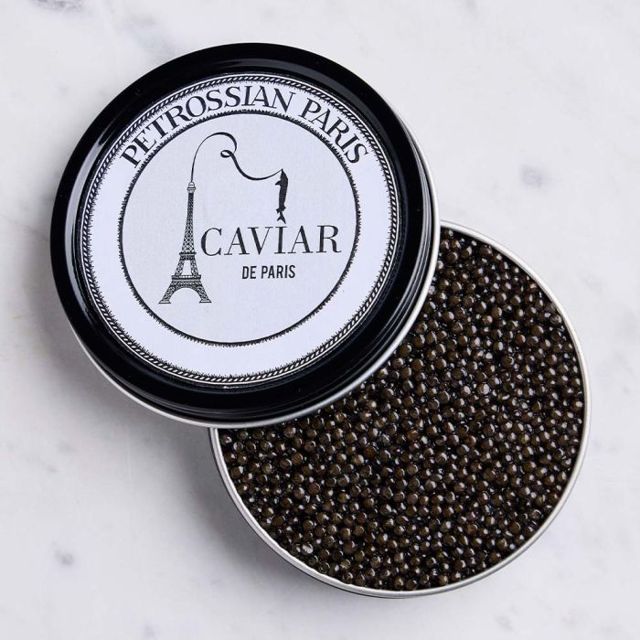 Une boîte ronde de caviar Petrossian Paris dont le couvercle est partiellement ouvert, révélant des perles de caviar noires à l'intérieur. Le couvercle, conçu par Claire Duclert, arbore le logo Petrossian avec une illustration de la Tour Eiffel et le texte « Caviar de Paris ». La boîte repose sur une surface en marbre.
